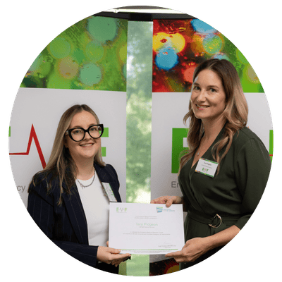 A circle image of two women standing indoors, one presenting a certificate to the other in front of a colourful backdrop featuring green and red elements with text.
