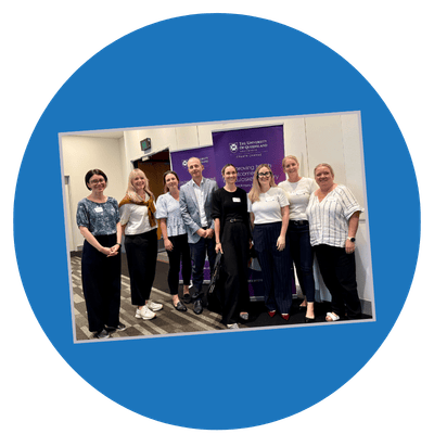 A photo of eight people standing indoors in front of a purple banner with University of Queensland branding. The landscape image is on a blue circle background titled slightly.