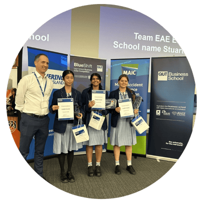 A circle image of three female students in school uniforms holding certificates and gift bags, standing next to a man in business attire at an award event. Background includes banners from QUT Business School and MAIC