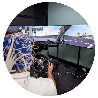 Person seated at a driving simulator wearing a cap fitted with multiple wires and sensors, holding a steering wheel. Three large curved monitors in front display a road intersection with traffic lights and lane markings. The simulator setup is in an indoor room with desks, cables, and equipment visible around the screens.