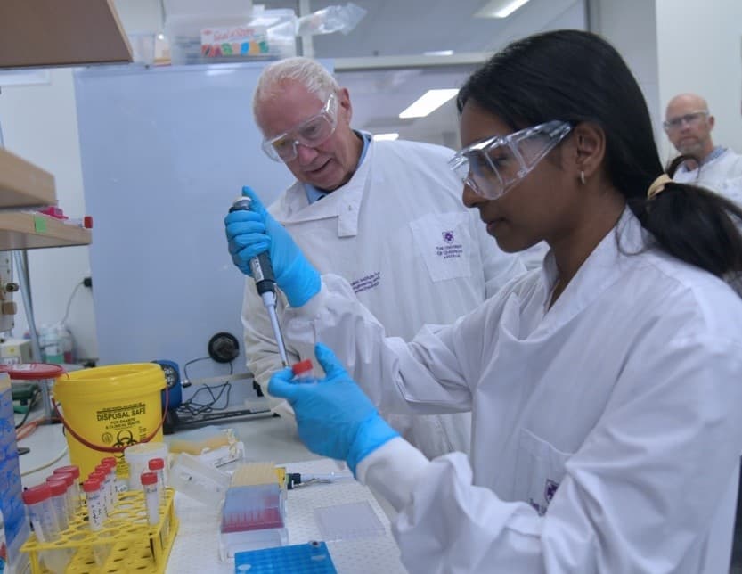 A woman wearing protective clothing and goggles uses scientific lab equipment as a man wearing similar watches.