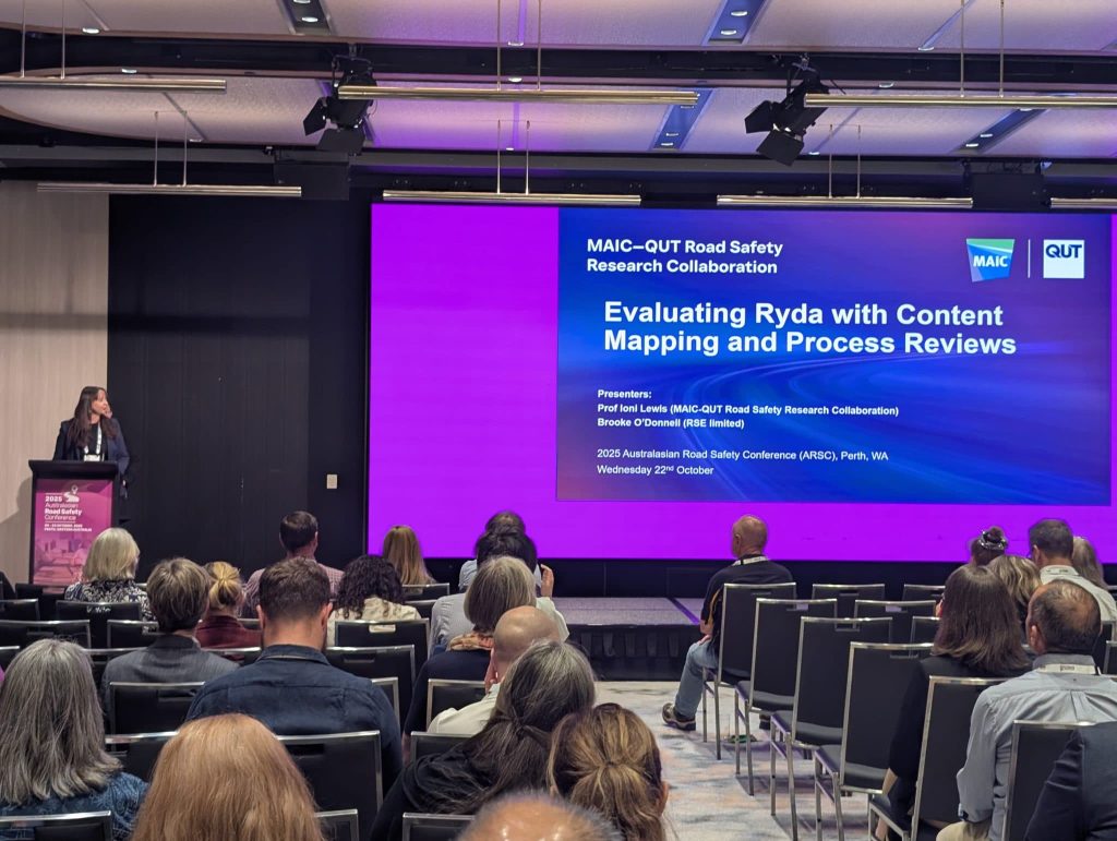 A women stands at a podium addressing a seated crowd. She looks to the right at a screen that reads, “MAIC-QUT Road Safety Research Collaboration Evaluating Ryda with Content Mapping and Process Reviews.”