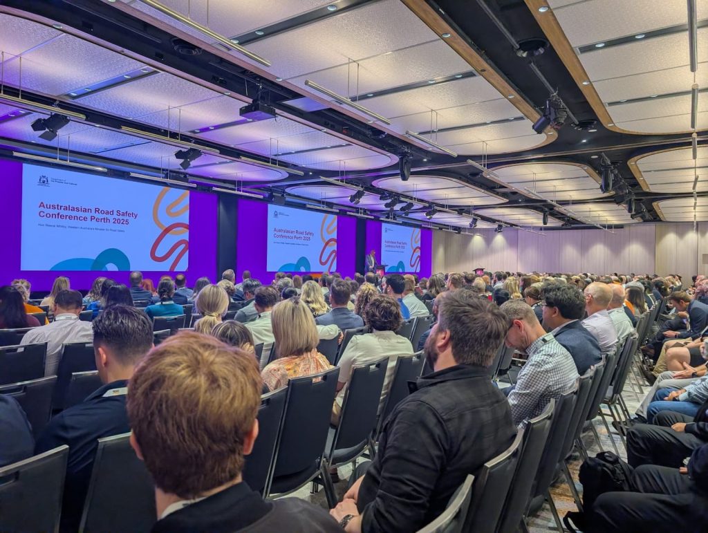 A room full of seated people look towards the front of the room to large screens that display “Australasian Road Safety Conference Perth 2025” with colourful geometric lines.