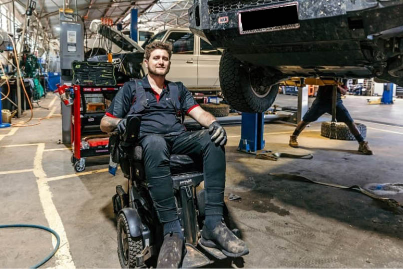 A smiling man in a wheelchair, wears all black work clothing. A mechanic’s workshop is visible behind him.