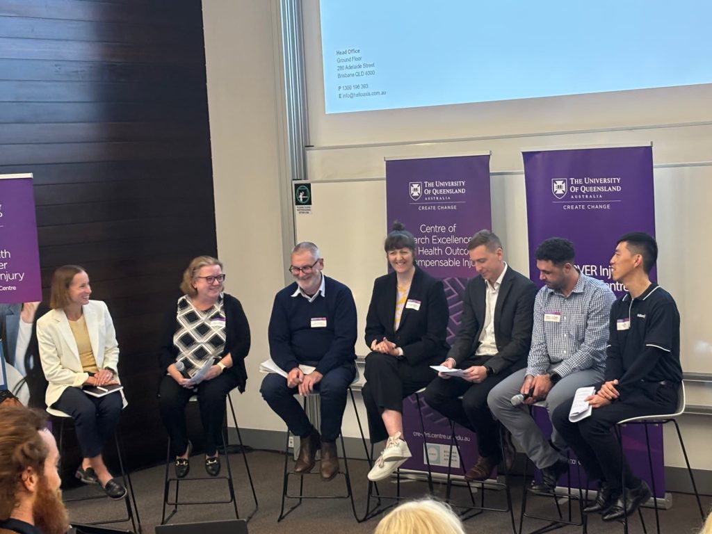 A group of seven people sit on chairs in a semi-circle. Behind them purple banners with “University of Queensland” logos are visible. Behind the banners is a screen displaying the text “Panel: Barriers to research implementation in the CTP insurance space.”