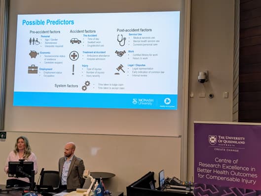 A man and woman stand behind a lectern, to their right a purple banner with the text “The University of Queensland Centre of Research Excellence in Better Health Outcomes for Compensable Injury” is visible. Above a PowerPoint presentation titled “Possible Predictors” is visible.