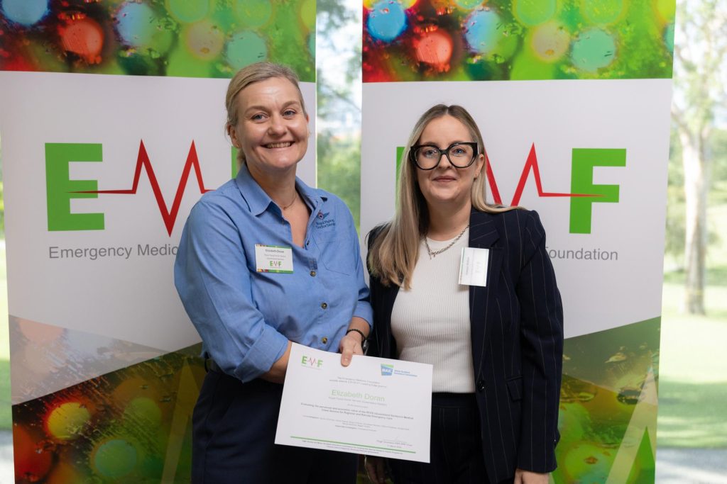 An image of two women standing in front of two green and red banners displaying "EMF Emergency Medicine Foundation”. One woman is showing a paper certificate with the EMF and MAIC logos on it.