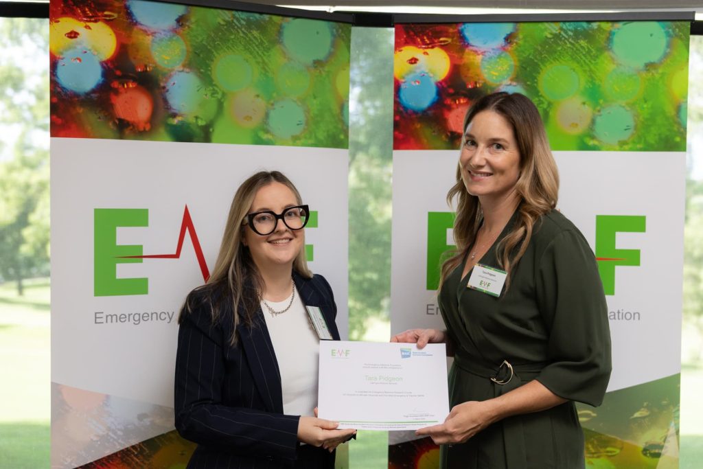 An image of two women standing in front of two green and red banners displaying "EMF Emergency Medicine Foundation”. Both women are smiling, with one handing a paper certificate to the other.
