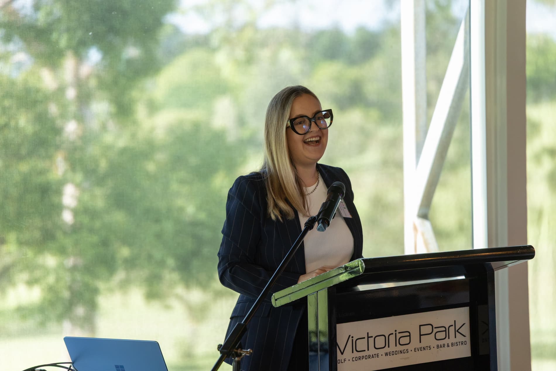 An image of a woman standing behind a podium labelled "Victoria Park," delivering a speech with a microphone. The background is a large window with a blurred view of green.