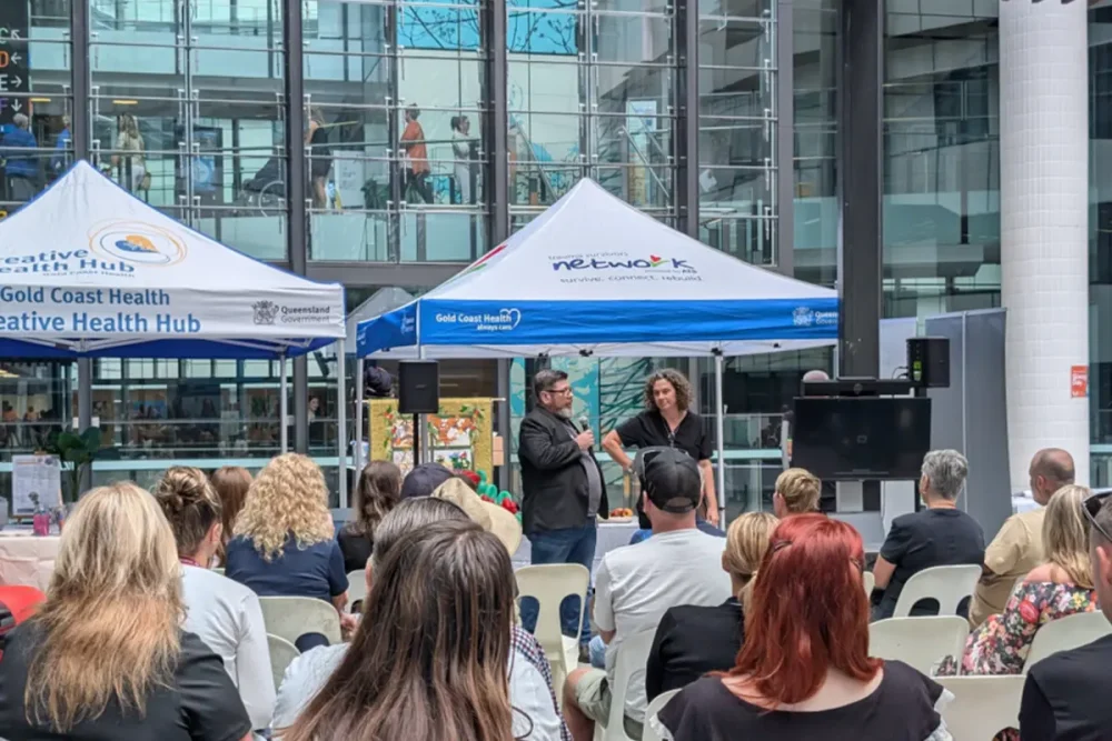 An image of a large audience from behind, sitting in chairs outside a building with large windows. They are facing two people standing and speaking with a microphone under a tent labelled “tauma suvivors network”.