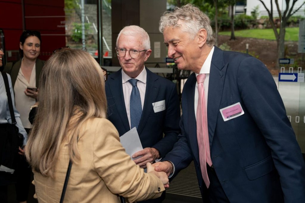 An image showing a professional networking event where two men in suits and a woman in a beige jacket are engaged in a handshake and conversation.