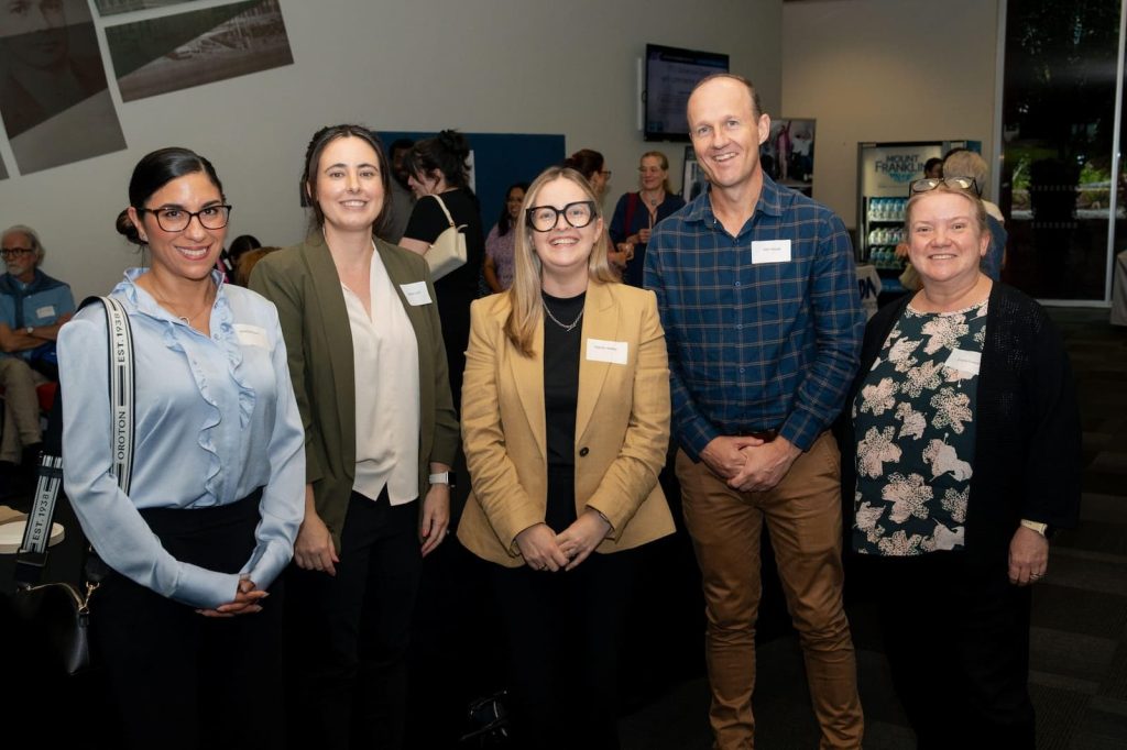 An image of four women and one man standing together in a row, smiling at the camera. They are dressed in business casual attired with name tags on, and other attendees are seen networking in the background.