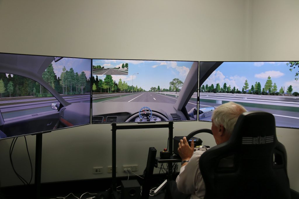 View of man in driving simulator. Only the back of his head, hands and arms are visible. He is seated and operating a steering wheel. Three large screens displaying road scenery are visible. He is in a room with white walls.