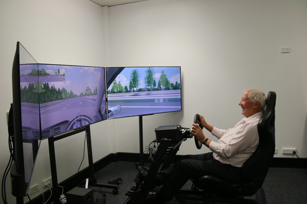 Side view of a smiling man in driving simulator. He is seated and operating a steering wheel. Three large screens displaying road scenery are visible. He is in a room with white walls.