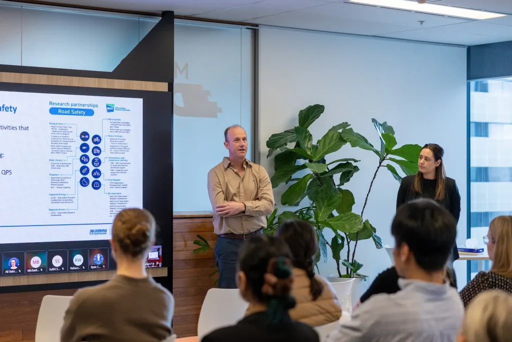 A man speaks to attendees during a Queensland Road Safety Week session, with road safety research content displayed on a large screen behind him.