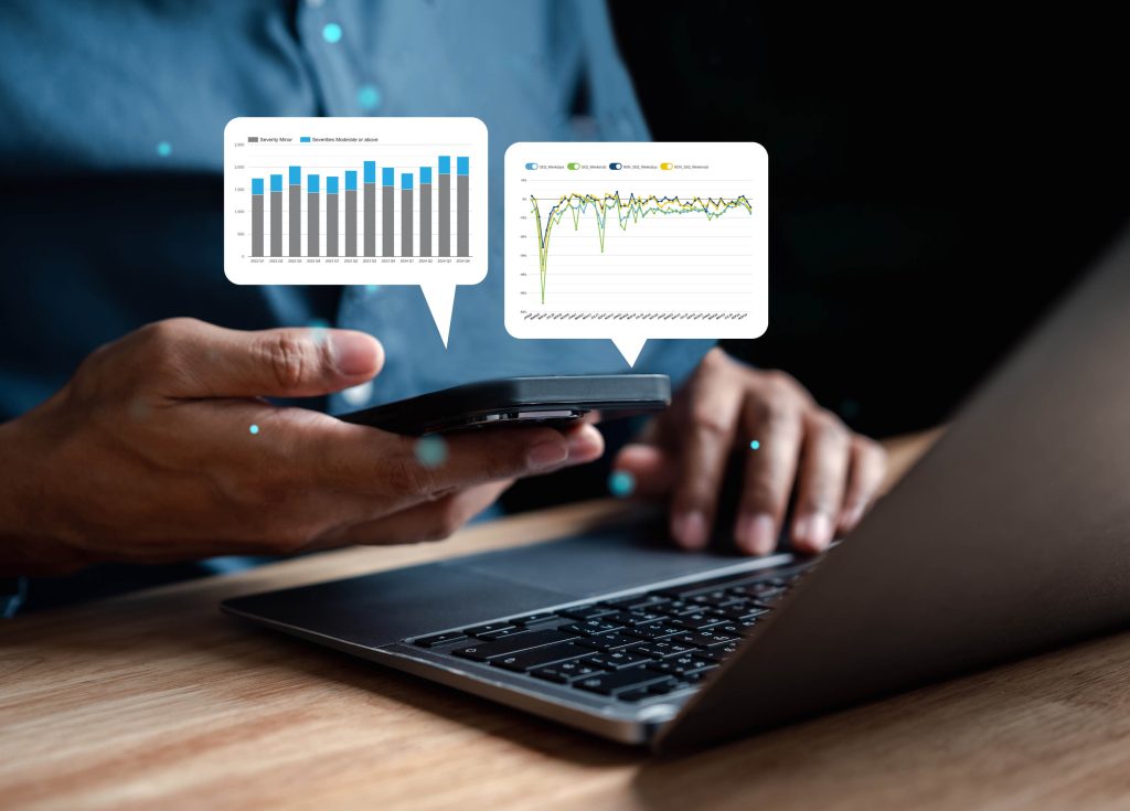 Hands hold a mobile phone in front of a person’s chest wearing a blue button up shit. Speech bubbles with graphs in them sit above the phone. A laptop is laid out on the desk in front.