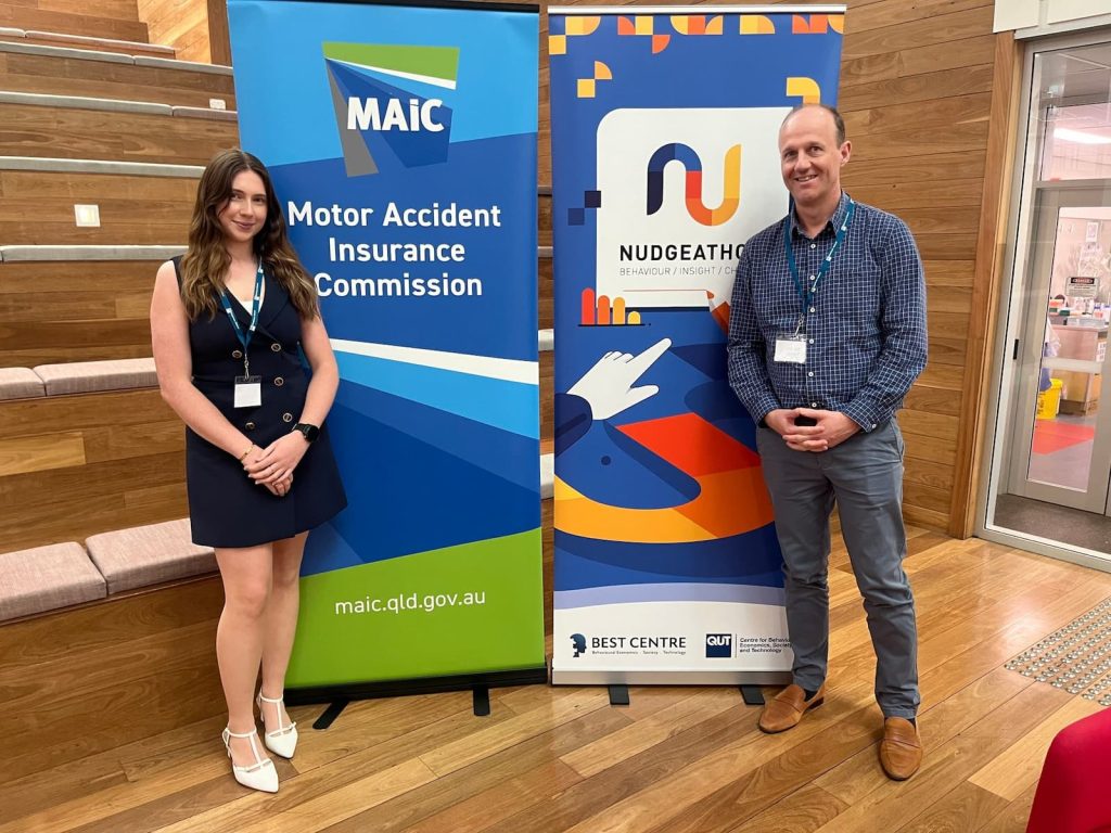 A smiling man and woman stand either side of two pull-up banners. One banner reads “MAIC Motor Accident Insurance Commission” the other banner reads “Nudgeathon.”