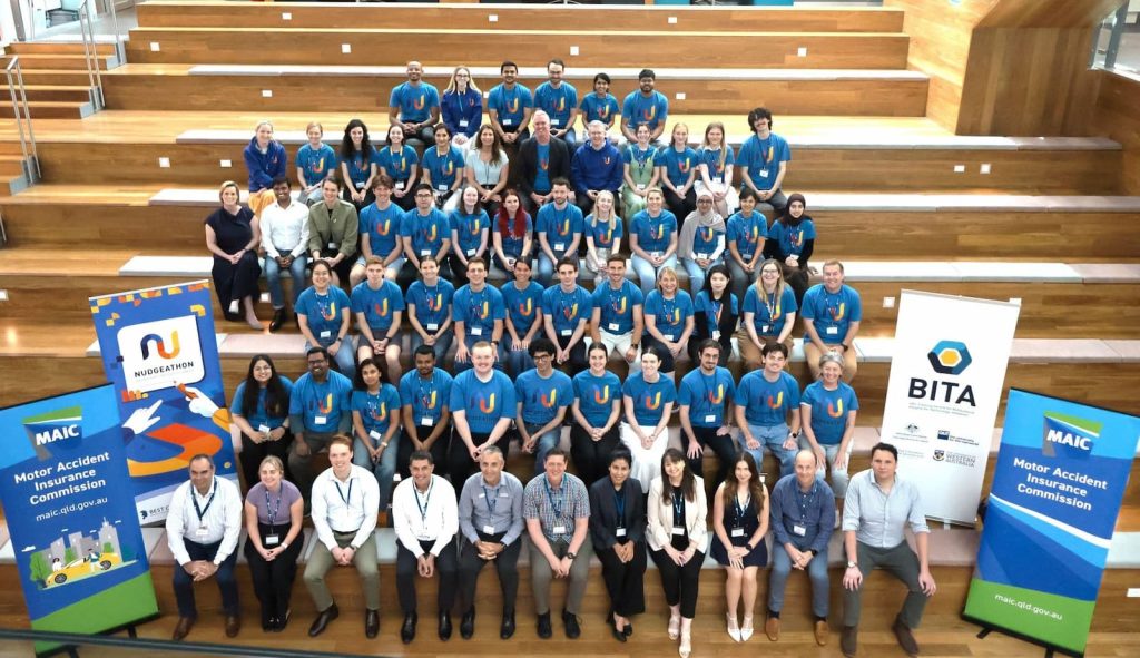 A large crowd of people are seated on stairs. Many are wearing blue t-shirts. Four banners are either side of the group. Two of the banners read Motor Accident Insurance Commission. The other two read Nudgeathon and BITA.