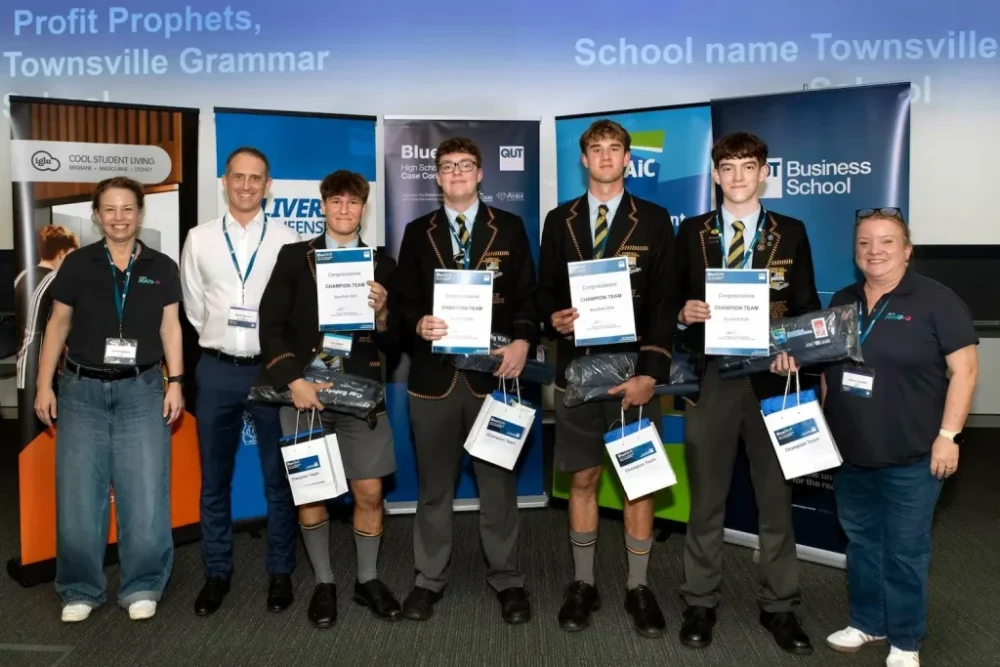 Two female and one male judge from MAIC posing with the four students from the winning team for BlueShift 2026, standing in front of various pull up banners promoting key sponsors of the event.