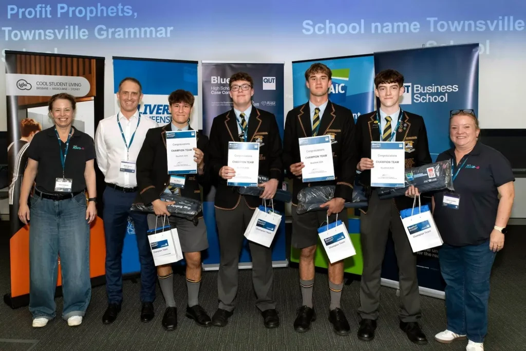 Two female and one male judge from MAIC posing with the four students from the winning team for BlueShift 2026, standing in front of various pull up banners promoting key sponsors of the event.