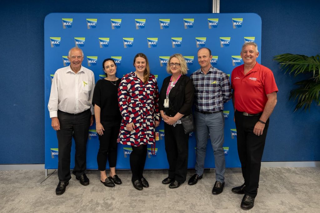 Six smiling people stand in front of a blue media wall with “MAIC” logos.