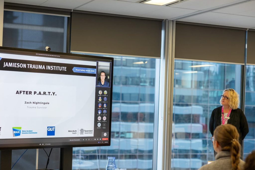 A woman stands staring at a large screen. On the screen is text that reads “Jamieson Trauma Institute. After P.A.R.T.Y. Zach Nightingale.