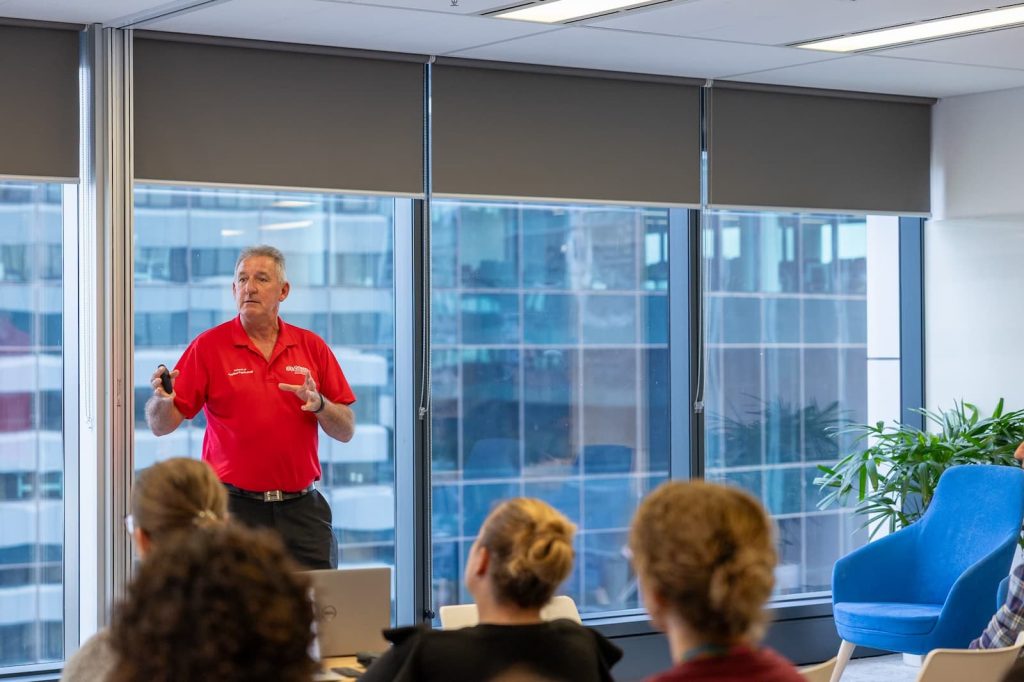 A man wearing a red shirt holds his hands out as he stands in front of a room of seated people, windows and buildings are visible behind him.