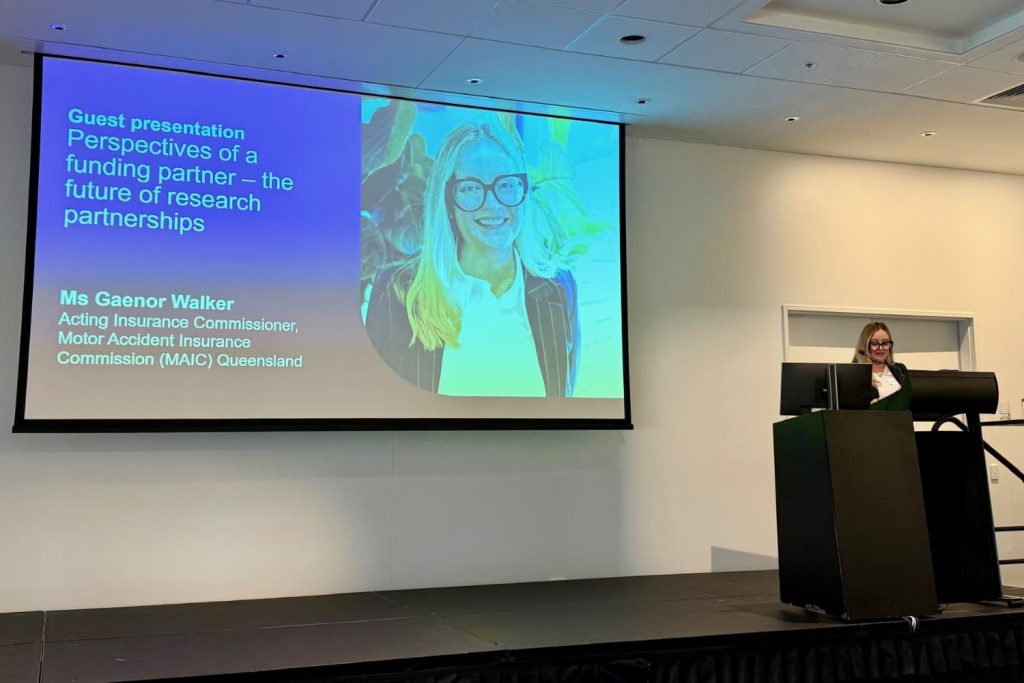 A photograph of a conference presentation showing a speaker standing at a podium beside a large screen displaying a slide titled "Guest presentation: Perspectives of a funding partner – the future of research partnerships." Slide includes presenter name Ms Gaenor Walker, Acting Insurance Commissioner at Motor Accident Insurance Commission (MAIC) Queensland, with a photo of the presenter.