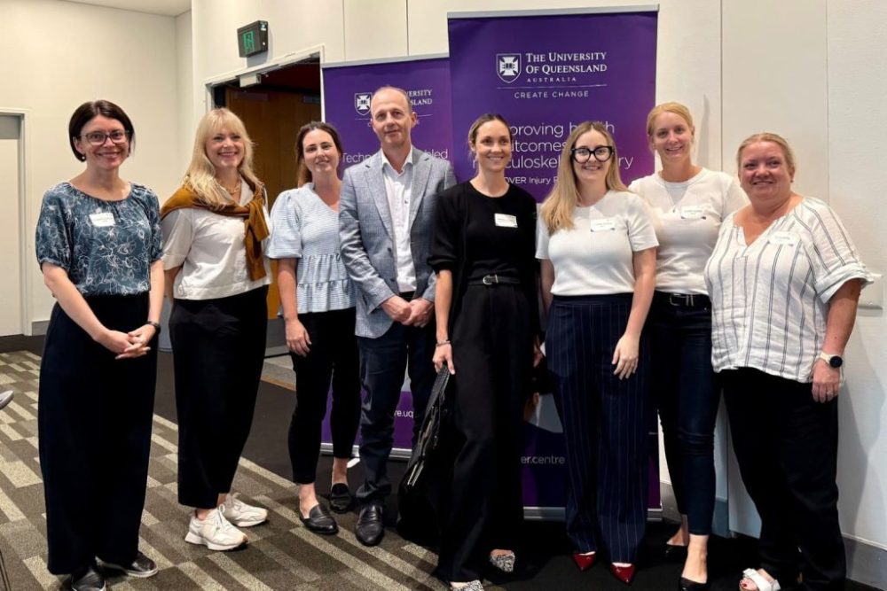 Eight smiling people stand in a row. University of Queensland banners are visible behind them.