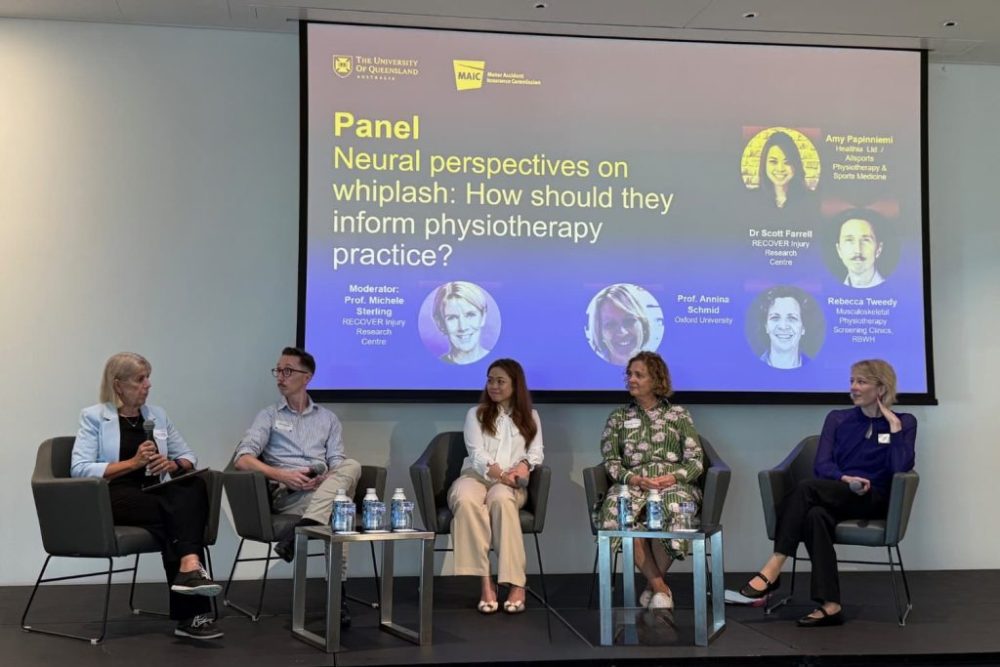 A photograph of a panel discussion titled "Neural perspectives on whiplash: How should they inform physiotherapy practice?" featuring five seated speakers on stage with a large presentation slide behind them. Slide includes names, roles, and photos of panellists, with white text on a dark blue background, highlighting experts from University of Queensland and related institutions discussing physiotherapy approaches to whiplash.
