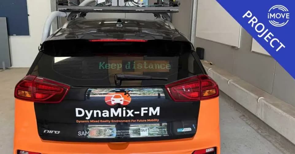 Close up of the back of a black and organge car. There is white text on the car that reads “DynaMix-FM” and text on the back windscreen that reads “Keep distance”. In the corner of the image is a blue triangle with white text that reads “iMOVE PROJECT