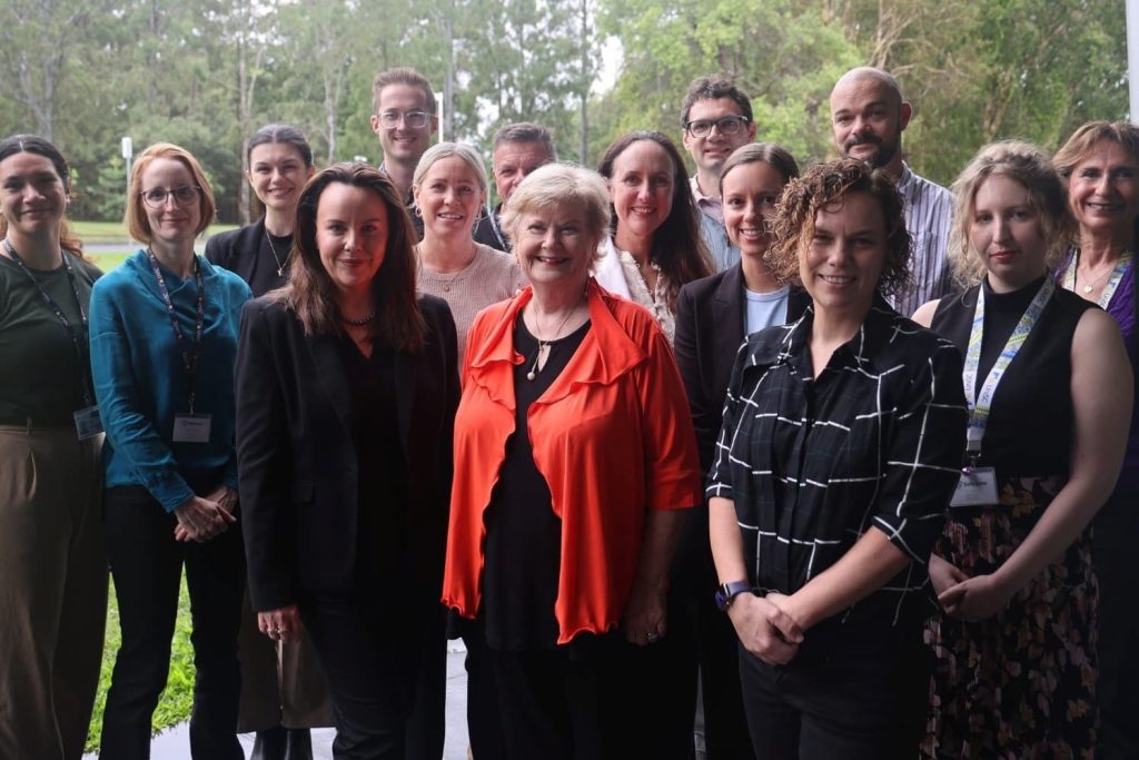 Photograph of a group of 15 people standing outdoors in front of greenery, posing for a large group photo