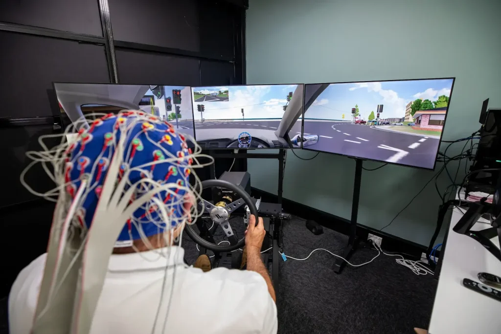 A person wearing a blue cap with numerous wires extending from it operates a driving simulator